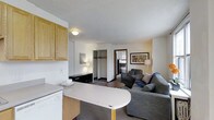This single-floor unit presents an open-concept layout that seamlessly connects the kitchen and living areas, creating an efficient and practical living space. The kitchen is outfitted with light oak cabinetry providing ample storage, a white countertop that extends into a peninsula-style breakfast bar, and standard appliances including a dishwasher. The textured white ceiling and neutral wall tones lend a clean, understated aesthetic throughout.

The living area is carpeted and comfortably accommodates a gray upholstered sofa and armchair arranged around a round coffee table, with framed artwork adorning the walls and floor lamps providing warm ambient lighting. A recessed closet with a hanging rod offers built-in storage and is centrally visible from the main living space.

A doorway leads to a separate room beyond the living area, suggesting additional functional space such as a bedroom or den. Natural light filters in through a window visible in the background, contributing to the overall brightness of the unit. The layout is straightforward and well-suited for efficient everyday living.