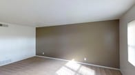 This two-story space provides a practical and understated layout designed for comfortable everyday living. The interior is characterized by wall-to-wall carpeting that adds warmth underfoot, complemented by clean white baseboards that frame each room with a crisp finish. The walls are painted in earthy, neutral tones — as seen in the deep olive-brown accent wall visible in one of the main rooms — creating a grounded, cozy atmosphere throughout. Textured popcorn ceilings are consistent across the space, adding a classic touch to the overall aesthetic. Natural light filters generously into the rooms, casting soft, dynamic patterns across the carpeted floors and contributing to a bright and welcoming ambiance. Electrical outlets are conveniently positioned along the walls, reflecting a thoughtfully functional design. Spanning two floors, the space offers separation between living areas, providing distinct zones that can serve a variety of purposes. The overall character of the space is clean, neutral, and move-in ready, making it well-suited for those looking for a straightforward and livable environment with room to personalize.