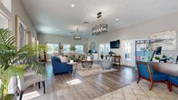 Located at 13125 Wilcox Road in Largo, Florida, this thoughtfully designed single-floor community or leasing office offers a generously proportioned open-plan interior that seamlessly blends functionality with inviting, coastal-inspired style. The space is defined by soaring ceilings that amplify its sense of openness, while an abundance of natural light streams in through multiple large windows and sets of French doors fitted with transom windows above — illuminating the warm neutral tones of the walls throughout.

The entry foyer features classic large-format tile flooring and elegant wingback seating, creating a polished first impression. Moving deeper into the space, the flooring transitions to rich wood-look hardwood, anchoring distinct zones that include a comfortable lounge area with a bold navy sofa, upholstered accent chairs, and layered area rugs. Multiple wire-cage pendant chandeliers hang from the ceiling, adding an industrial-chic touch that complements the coastal aesthetic.

A built-in workstation with stone-topped cabinetry and dual computer monitors serves as a practical business hub, accented by a striking oversized metal wall clock. A reception/leasing desk with blue upholstered guest chairs provides a dedicated service area, while a wall-mounted flat-screen TV and French doors opening to an outdoor patio round out the amenities. Lush potted greenery and curated artwork complete the welcoming ambiance.