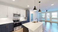 This single-floor, 2-bedroom, 2-bathroom residence offers a thoughtfully designed layout with modern finishes throughout. The open-concept kitchen serves as the centerpiece of the living area, showcasing a striking two-tone cabinetry scheme — crisp white upper cabinets paired with deep navy-blue lower cabinets — all accented with gold and brushed nickel hardware. A central island with a white quartz countertop, undermount sink, and matte black faucet anchors the space, illuminated by a pair of industrial-style pendant lights. Stainless steel appliances, including a refrigerator, electric range, and built-in microwave, complete the kitchen. The herringbone-patterned hardwood flooring in the main living area adds a distinctive architectural touch, while recessed lighting ensures a bright, welcoming atmosphere. Both bedrooms feature plush carpeting and large windows fitted with blinds that invite natural light. The primary bedroom includes a walk-in closet with multi-tier shelving and hanging rods. The primary bathroom features a soaking tub with bold hexagonal dark-gray tile surround and wood-look tile flooring. A hallway and two dedicated closets provide practical storage solutions throughout the residence.