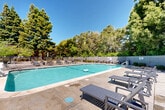 Located at 3500 Edison Street in San Mateo, California, this outdoor amenity area centers around a generously sized rectangular swimming pool with clear, well-maintained turquoise water. The pool is finished with a smooth concrete surround that extends into a broad sundeck, providing ample space for relaxation and recreation. Numerous lounge chairs and seating arrangements are distributed around the deck, offering residents plenty of options for sunbathing or casual outdoor gatherings. A dedicated hot tub or spa feature is positioned adjacent to the main pool, adding to the versatility of the outdoor space. The area is enclosed by a perimeter fence and framed by a lush canopy of mature trees — including tall redwoods and broad deciduous trees — that provide natural shade, privacy, and a serene, park-like atmosphere. A pool house or utility structure is situated at one end of the deck, offering practical support facilities. The combination of open sky exposure and surrounding greenery creates a bright yet sheltered environment well-suited for year-round outdoor enjoyment in the mild San Mateo climate. The overall setting is clean, well-kept, and thoughtfully arranged to maximize both comfort and functionality.