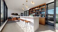 Located at 11311 La Grange in Los Angeles, CA 90025, this single-story space showcases a compelling blend of modern industrial and mid-century design elements. The entry lobby immediately impresses with its polished concrete floors that reflect abundant natural light streaming through floor-to-ceiling glass sliding doors. A dramatic board-formed concrete accent wall anchors the entry, complemented by a sleek decorative rock bed at its base, lending an organic, architectural contrast to the otherwise clean, white surroundings.

The interior transitions seamlessly into a sophisticated lounge and bar area, where a ribbed white bar island takes center stage, lined with sculptural wood-and-metal barstools. Overhead, elegant brass pendant lights with globe shades illuminate the space warmly. A full wall of open-faced, warm-toned wood shelving provides both display storage and visual depth, showcasing curated décor pieces. An oval mirror and additional wood cabinetry further reinforce the mid-century modern aesthetic throughout.

Recessed lighting, crisp white ceilings, and the consistent use of polished concrete flooring unify the space, creating an open, airy ambiance. The thoughtful interplay of natural textures — raw concrete, warm wood, and metal accents — gives this property a refined yet approachable character well-suited for a variety of uses.