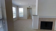 This single-story space showcases a thoughtfully designed open-concept living area finished in a neutral, light color palette throughout. The main living room is carpeted in a warm beige tone and benefits from natural light streaming through multiple windows, offering views of surrounding greenery. Crown molding adds a refined architectural detail along the ceiling, contributing to a clean, polished aesthetic.

A tile-surround fireplace anchors one side of the living area, providing both a functional and visual focal point. Adjacent to the fireplace, a white-painted half-wall with a dark countertop top serves as a room divider, subtly separating the living space from a neighboring area while maintaining an open feel. HVAC vents are integrated into the ceiling, ensuring efficient climate control throughout.

A centrally located hallway leads further into the unit, hinting at additional rooms beyond. White paneled doors add consistency to the interior design. The overall ambiance is calm and move-in ready, with a neutral finish palette that offers flexibility for a variety of furnishing styles.