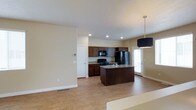 This two-story home is designed with an open and functional layout that maximizes the sense of space throughout. The main floor showcases an open-concept living and kitchen area bathed in natural light, with light-toned hardwood-style flooring that extends seamlessly throughout the space.

The kitchen is a standout feature, equipped with rich dark wood cabinetry, granite countertops, and a full suite of black appliances including a gas range, microwave, and refrigerator. A central island with an integrated sink serves as both a practical workspace and a gathering point, accented by a stylish pendant light overhead. Recessed lighting in the ceiling adds a clean, modern touch to the kitchen area.

The open layout connects the kitchen effortlessly to the living and dining areas, creating a spacious and inviting atmosphere ideal for everyday living and entertaining. Neutral warm tones on the walls complement the overall aesthetic, giving the space a bright and welcoming feel. A door near the kitchen provides convenient access to the exterior, allowing natural light to filter in.

The two-story design offers additional living space on the upper level, providing separation between common areas and private quarters. Overall, the home presents a well-balanced combination of modern finishes and practical design throughout.