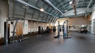 Located at 120 Perry St in Petersburg, VA, this fitness facility offers a generously sized, single-floor workout space defined by its distinctive architectural character. The most striking feature is the curved, barrel-vault metal roof supported by arched steel ribs, giving the space an industrial yet dynamic aesthetic. Exposed ductwork runs along the ceiling, complementing the raw, utilitarian design while pendant lighting provides even illumination throughout the floor.

The workout area is finished with durable black rubber flooring suited for heavy use and is divided functionally into strength and cardio zones. The strength section houses a variety of selectorized machines, including cable systems, a leg press, and additional resistance equipment, along with free weights such as dumbbells. A wall-length mirror along one side enhances the sense of space and allows for form monitoring during training.

The cardio zone features treadmills, stationary bikes, a rowing machine, and other aerobic equipment positioned near large floor-to-ceiling glass windows and doors. These glazed partitions allow natural light to filter in and offer views into adjacent areas of the building, including a staircase, creating a sense of connectivity. The open layout and soaring ceiling height provide an airy, motivating environment well-suited for a full-service fitness facility.