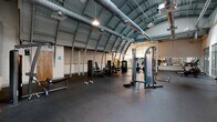 Located at 120 Perry St in Petersburg, VA, this fitness facility offers a generously sized, single-floor workout space defined by its distinctive architectural character. The most striking feature is the curved, barrel-vault metal roof supported by arched steel ribs, giving the space an industrial yet dynamic aesthetic. Exposed ductwork runs along the ceiling, complementing the raw, utilitarian design while pendant lighting provides even illumination throughout the floor.

The workout area is finished with durable black rubber flooring suited for heavy use and is divided functionally into strength and cardio zones. The strength section houses a variety of selectorized machines, including cable systems, a leg press, and additional resistance equipment, along with free weights such as dumbbells. A wall-length mirror along one side enhances the sense of space and allows for form monitoring during training.

The cardio zone features treadmills, stationary bikes, a rowing machine, and other aerobic equipment positioned near large floor-to-ceiling glass windows and doors. These glazed partitions allow natural light to filter in and offer views into adjacent areas of the building, including a staircase, creating a sense of connectivity. The open layout and soaring ceiling height provide an airy, motivating environment well-suited for a full-service fitness facility.
