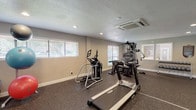 This fitness center is a comprehensively equipped, single-floor exercise facility designed to support a full range of workout routines. The space features durable rubber flooring throughout, providing a safe and resilient surface ideal for high-impact activity and heavy equipment. Recessed lighting is complemented by multiple windows fitted with horizontal blinds, allowing ample natural light to fill the room and create a bright, energizing atmosphere. A ceiling-mounted HVAC unit ensures comfortable climate control year-round.

The facility is outfitted with a diverse selection of equipment to accommodate both cardiovascular and strength training needs. Cardio options include a treadmill, an elliptical trainer, and a stationary exercise bike. For strength training, a multi-station cable machine offers a variety of resistance exercises, while a two-tier dumbbell rack stocked with a full range of free weights supports targeted muscle work. Assorted exercise balls add versatility for balance and core conditioning.

A large wall mirror spans one side of the room, visually expanding the space and allowing users to monitor their form during workouts. A posted fitness center rules sign reflects an organized, community-oriented environment. The space is accessed via a glass-panel exterior door, with an emergency exit also present for safety compliance.