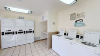 This shared laundry facility is designed with functionality and convenience in mind. The room features light beige walls accented by crown molding, and clean, light-toned tile flooring that creates a bright and welcoming atmosphere. 

The space is outfitted with multiple top-loading washing machines neatly arranged along one wall, complemented by a bank of stacked dryers on the opposite side, providing ample capacity for residents. A built-in shelving unit with a folding or sorting ledge is centrally positioned, offering a practical surface for handling laundry between cycles.

Ventilation is well-considered, with visible wall vents ensuring proper airflow throughout the room. Overhead lighting keeps the space well-lit, while a decorative skylight-style ceiling fixture adds a subtle architectural touch. Cheerful laundry-themed wall signs contribute a lighthearted, welcoming character to the otherwise utilitarian space.

Overall, the facility is clean, organized, and thoughtfully laid out to accommodate multiple users simultaneously, making it a highly practical shared amenity.