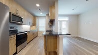 This single-floor space showcases a thoughtfully designed open-concept layout where the kitchen and living area flow seamlessly together. The kitchen is outfitted with flat-front wood-toned cabinetry that extends from floor to ceiling, providing ample storage. Dark granite countertops offer a striking contrast to the warm cabinetry, and a peninsula with an integrated sink creates a natural division between the kitchen and living area while also serving as additional workspace or casual seating. Stainless steel appliances, including a refrigerator with an ice dispenser, electric range, built-in microwave, and a textured tile backsplash complete the kitchen's modern aesthetic.

Light hardwood-style flooring runs continuously throughout the space, contributing to a clean, cohesive look. The open living area is bright and versatile, featuring neutral walls, a ceiling fan with a built-in light fixture, and large windows fitted with horizontal blinds that allow natural light to filter in. A set of glass double doors leads to the exterior, further enhancing the sense of openness and connectivity to the outdoors. Recessed lighting supplements the natural light, ensuring the space remains well-lit throughout the day.