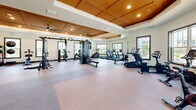 Located at 904 S Lake St in Leesburg, Florida, this well-appointed fitness center offers a generously sized, single-floor exercise space designed to support a comprehensive workout experience. The room's architectural design is immediately striking, showcasing elevated coffered ceilings with warm, honey-toned wood paneling and recessed lighting that creates a bright, inviting atmosphere. Large ceiling fans are strategically positioned throughout to ensure comfortable airflow across the open floor plan.

Natural light pours in through multiple windows along the perimeter walls, offering views of the surrounding outdoor environment and contributing to an energizing workout setting. Full-length mirrors line sections of the walls, enhancing the sense of openness while allowing users to monitor their form.

The exercise floor features durable, rubberized flooring designed for both strength and cardio activities. Equipment includes a robust selection of selectorized strength machines, cable systems, free weights, weight benches, a TRX suspension system, treadmills, rowing machines, stationary bikes, and elliptical trainers. A wall-mounted television provides entertainment during workouts. The thoughtful layout ensures ample spacing between equipment, allowing for a comfortable and safe training environment throughout the entire space.