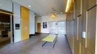 This building lobby presents a polished, contemporary aesthetic that makes an immediate impression upon entry. The space is defined by striking warm-toned wood paneling that lines the walls, creating a rich contrast against the crisp white walls and smooth large-format gray tile flooring. A statement pendant light fixture with an organic, starburst design hangs from the ceiling, adding an artistic focal point and a warm golden glow to the environment. Recessed ceiling lighting supplements the ambient illumination, ensuring the space feels bright and inviting throughout. Two stainless steel elevator doors anchor the far wall, their sleek metallic finish complementing the modern design language of the lobby. A minimalist bench with a yellow-green top and slim metal legs sits centrally in the space, offering a functional yet design-forward seating element for visitors. A framed black-and-white photograph adorns the wall, adding a subtle artistic touch. A glass partition wall is visible to one side, providing transparency while defining separate zones within the lobby. The overall ambiance is clean, professional, and thoughtfully curated, reflecting a high standard of contemporary interior design.