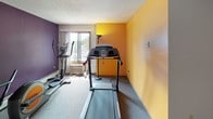 This fitness room offers a dedicated exercise space designed to support a variety of cardio-focused workouts. The room is immediately distinguished by its bold color scheme, featuring striking purple and golden yellow accent walls that create an energetic atmosphere against a neutral white ceiling finished with a textured surface.

The space is equipped with several pieces of cardio equipment, including a treadmill, an elliptical trainer, and a stationary exercise bike, providing users with multiple options for low- and high-impact workouts. A set of free weights is also available near the window area. Built-in sliding wood-panel storage closets line one wall, offering practical storage solutions within the room.

Carpeted flooring runs throughout, and a window along one wall allows natural light to filter in while offering a view to the outdoors. A wall-mounted air conditioning unit beneath the window helps maintain a comfortable environment during exercise. The room is accessed through a wood-trimmed door, beside which a posted sign outlines the exercise room rules. A folding chair provides a convenient rest area. Overall, the space is straightforward, functional, and well-equipped for everyday fitness needs.