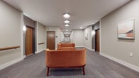 This single-floor corridor space is thoughtfully designed to balance functionality with comfort. The hallway extends generously, lined with rich wood-toned entry doors accented by darker door frames that add visual contrast against the neutral gray walls. Continuous wooden handrails run along both sides of the corridor, providing accessibility support throughout the length of the space.

At key intervals along the hallway, curated seating areas break up the linear flow, each featuring tufted burnt-orange velvet sofas and matching armchairs paired with a plaid upholstered ottoman. These furnishing choices introduce warmth and a residential feel to what might otherwise feel purely transitional. Framed abstract artwork in soft coral and blue tones is mounted along the walls, complementing the overall color palette.

The flooring consists of patterned low-pile carpet in muted gray-blue tones, contributing to both sound absorption and visual cohesion. Overhead, flush-mount circular light fixtures provide consistent illumination throughout, supplemented by recessed lighting and wall-mounted sconces near the entry doors. The clean white ceiling and baseboards provide crisp definition, giving the corridor a polished, well-maintained appearance throughout.