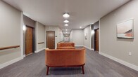 This single-floor corridor space is thoughtfully designed to balance functionality with comfort. The hallway extends generously, lined with rich wood-toned entry doors accented by darker door frames that add visual contrast against the neutral gray walls. Continuous wooden handrails run along both sides of the corridor, providing accessibility support throughout the length of the space.

At key intervals along the hallway, curated seating areas break up the linear flow, each featuring tufted burnt-orange velvet sofas and matching armchairs paired with a plaid upholstered ottoman. These furnishing choices introduce warmth and a residential feel to what might otherwise feel purely transitional. Framed abstract artwork in soft coral and blue tones is mounted along the walls, complementing the overall color palette.

The flooring consists of patterned low-pile carpet in muted gray-blue tones, contributing to both sound absorption and visual cohesion. Overhead, flush-mount circular light fixtures provide consistent illumination throughout, supplemented by recessed lighting and wall-mounted sconces near the entry doors. The clean white ceiling and baseboards provide crisp definition, giving the corridor a polished, well-maintained appearance throughout.