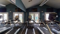 Located at 8301 Paces Oaks Blvd in Charlotte, NC, this single-floor fitness center is a well-equipped facility designed to support a comprehensive workout experience. The space is defined by its bold teal-painted walls, durable tiled flooring, and large wall mirrors that visually expand the room while allowing exercisers to monitor their form.

The cardio zone is generously appointed with multiple treadmills, elliptical machines, and a stationary bike, all positioned to take advantage of natural light streaming in through several large windows and glass French doors. Mounted flat-screen TVs are positioned above the cardio equipment for entertainment during workouts.

The strength training area features a cable cross machine, a dumbbell rack with a variety of free weights, medicine balls, and adjustable workout benches, providing ample options for resistance and functional training. A ceiling fan and overhead lighting ensure the space remains comfortable and well-lit throughout the day.

Natural light plays a significant role in the ambiance of this fitness center, with windows and glass doors offering views of an outdoor patio area with seating. The combination of practical equipment, mirrored walls, and a bright, open layout makes this fitness center a functional and inviting space for residents seeking a convenient on-site workout facility.