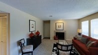 This single-floor space offers a well-appointed living area designed with both comfort and style in mind. The living room serves as the focal point of the home, centered around a tile-surround fireplace that adds warmth and character to the space. Neutral beige walls and carpeted flooring create a calm, inviting backdrop throughout.

The living room is furnished with a rich, rust-toned sofa and coordinating accent chairs, complemented by a sleek glass-top coffee table that adds a contemporary touch. A bold black-and-white floral area rug anchors the seating arrangement, providing a striking contrast to the soft carpeting. Decorative red ceramic accents and framed wall art add pops of color and personality to the room.

A low-profile black sideboard with vertical slat detailing lines one wall, topped with decorative vases and sculptures that enhance the space's curated aesthetic. A HVAC ceiling vent ensures year-round climate comfort, while recessed lighting and natural light contribute to the overall brightness of the room. A hallway visible from the living area leads to additional rooms, suggesting a functional and well-connected floor plan. The textured ceiling adds a classic finish to this warm and welcoming living environment.