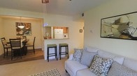 This single-story residence is designed with an open-concept floor plan that allows the living, dining, and kitchen areas to flow naturally into one another. The living area features light-colored carpeting and neutral wall tones, contributing to a bright and airy ambiance. A decorative pendant light adds a subtle accent overhead, while framed artwork on the walls adds a personal touch to the space.

The kitchen is accessible via a pass-through counter that doubles as a breakfast bar, accommodating two barstools with green cushioned seats. The kitchen itself is equipped with wood-toned cabinetry and a white refrigerator, visible through the open pass-through. This design element enhances connectivity between the kitchen and living spaces while maintaining a sense of defined zones.

Adjacent to the living area, a glimpse into the dining space reveals hardwood-style flooring and a dining chair, suggesting a comfortable area for meals. The overall color palette throughout the space is warm and neutral, with soft lighting enhancing the welcoming atmosphere. The layout is practical and well-organized, making efficient use of the available space.