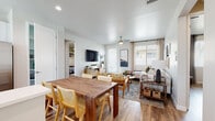 This thoughtfully designed single-floor residence offers a seamless blend of functionality and contemporary style throughout its open, light-filled layout. The combined living and dining area serves as the heart of the home, where warm wood-look flooring, recessed lighting, and a ceiling fan create a welcoming atmosphere. Large windows dressed with blinds and curtains allow natural light to pour in, while a wall-mounted television and dedicated media console anchor the living space. The dining area accommodates a solid wood table with rattan-accented chairs, positioned conveniently adjacent to the kitchen.

The kitchen is well-equipped with two-tone cabinetry—white upper cabinets paired with gray lower units—topped with clean white countertops. A peninsula provides additional prep space and casual seating, while stainless steel appliances, including a refrigerator, range, microwave, and dishwasher, complete the space.

The primary bedroom is bright and airy, featuring a large window and ample room for a queen-sized bed. A second bedroom is cleverly outfitted with a built-in Murphy bed system incorporating integrated shelving and storage, maximizing versatility. The full bathroom offers a shower-tub combination, a gray vanity with white countertop, and wood-look flooring, maintaining the cohesive aesthetic found throughout the residence.