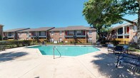 This single-story residence is part of a well-established brick exterior community, offering access to a generously sized outdoor swimming pool surrounded by a concrete deck and wrought iron fencing. Mature trees provide natural shade, adding to the relaxed outdoor ambiance under open skies.

Inside, the interior reflects a contemporary aesthetic built on a neutral foundation. The walls are finished in crisp white with a subtle texture, creating a clean and gallery-like atmosphere throughout. Polished concrete floors run continuously across the living areas, lending an industrial yet refined character to the space.

The dining area serves as a focal point, anchored by a striking round green marble table paired with woven rattan chairs atop a natural fiber rug. A large framed abstract artwork adorns the wall, reinforcing the curated, artistic sensibility of the interior. A sleek sliding barn door connects adjoining spaces, adding both functionality and a modern design detail.

Recessed lighting illuminates the interior evenly, while visible HVAC vents maintain a practical, well-ventilated environment. Glimpses into adjacent rooms reveal additional wall art and track lighting, suggesting a thoughtfully appointed and cohesive living space throughout.