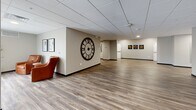 The lobby and lounge at 801 Kraewood Dr in Northfield, Minnesota, offers a generously proportioned, single-floor space designed to welcome and accommodate visitors with both functionality and style. The interior features durable wood-look plank flooring that runs throughout, complemented by a suspended tile ceiling with recessed and chandelier lighting that creates a warm, well-lit environment.

The lounge area serves as the centerpiece of the space, furnished with leather accent chairs in rich cognac and charcoal tones, a round dark-toned coffee table, and a plush area rug, all arranged to encourage comfortable conversation and relaxation. A dramatic oversized wall clock serves as a focal point on one of the neutral-toned accent walls, while framed artwork is thoughtfully displayed throughout, adding visual interest and character.

Toward the rear, a sleek reception desk is set against a textured dark wall panel, providing a professional and polished check-in area. Adjacent to the desk, a built-in credenza offers additional functionality, ideal for refreshment or service needs. Structural columns punctuate the open floor plan, adding architectural definition without disrupting the flow of the space. A glass entry door at one end allows natural light to filter in, connecting the interior to the outside. The overall ambiance is modern, clean, and professional.
