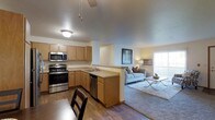 This single-floor space showcases an open-concept design that seamlessly connects the kitchen and living area, creating a cohesive and functional layout. The kitchen is well-appointed with stainless steel appliances, including a range, microwave, and dishwasher, complemented by warm honey-toned cabinetry that provides ample storage. Light-colored countertops run along the perimeter and extend to a peninsula, which serves as a natural divider between the kitchen and the adjoining living space while offering additional workspace. The flooring throughout is hardwood-style, lending warmth and continuity across both areas. A dining area is positioned near the kitchen, accommodating casual everyday meals. The living area is generously sized, featuring a large area rug, a sofa, and a glass-top coffee table, all beneath a vaulted ceiling that enhances the sense of openness and airiness. A window along the far wall allows natural light to filter into the living space, brightening the overall ambiance. The neutral color palette of whites, beiges, and warm browns throughout contributes to a calm and inviting atmosphere, making the space feel both practical and comfortable.