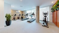 Located at 5630 Rue De Marseille in Montréal, this fitness facility offers a comprehensive workout environment across a generously sized single floor. The main exercise room is bright and modern, bathed in natural light through large floor-to-ceiling windows that frame views of the surrounding cityscape. Exposed concrete columns add an industrial architectural character, complementing the clean white walls and light-toned flooring throughout.

The cardio zone features multiple treadmills, elliptical trainers, and stationary cycling bikes arranged to maximize space and usability. A vibrant orange partition wall with integrated live plant displays serves as a stylish divider, separating the cardio area from the strength training section. The strength zone is well-appointed with a multi-function cable machine, a power rack, free weights, adjustable benches, and built-in shelving units with warm wood accents for organized equipment storage.

A dedicated stretching and functional training area features mirrored walls, light wood cabinetry, resistance bands, and yoga mats, creating a calm and focused environment for flexibility work. Recessed lighting throughout ensures consistent, comfortable illumination. The facility also includes a practical bathroom finished with terrazzo-style surfaces and bold red accent walls, equipped with accessibility grab bars. Overall, the space strikes a balance between functionality and contemporary design.
