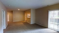 This single-floor residential unit offers a clean, functional layout with a welcoming ambiance defined by neutral warm tones and understated finishes throughout. The main living area is spacious and open, featuring wall-to-wall plush carpeting that adds comfort underfoot and a sense of warmth to the room. Smooth, white ceilings with recessed lighting provide even illumination, while the simple, unadorned walls offer a blank canvas ready for personalization.

A pass-through opening connects the living area to the kitchen, creating an accessible and communicative flow between spaces. The kitchen is efficiently designed with light wood cabinetry providing ample storage, a countertop that extends to form a convenient bar-style serving ledge, and standard white appliances including a microwave and range. The kitchen area features hard flooring, contrasting with the carpeted living space and helping to delineate the two zones.

Clean white baseboards frame each room, adding a crisp finishing detail against the warm beige walls. The overall layout is practical and well-proportioned, making efficient use of the available space. The unit's straightforward design and neutral palette make it highly adaptable to a variety of furnishing styles and personal tastes.