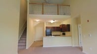 This two-story space is defined by its dramatic vertical design, with soaring double-height ceilings in the main living area that create an expansive, airy atmosphere. The open-concept layout seamlessly connects the living space to the kitchen, which features rich dark wood cabinetry, black appliances including a refrigerator and built-in microwave, and a peninsula-style breakfast bar with a light-toned countertop that provides both additional prep space and a casual dining option. Pendant lighting hangs above the kitchen area, complementing the flush-mount ceiling fixtures throughout. A staircase along one wall leads to the upper level, where a loft-style overlook is enclosed by a classic wood baluster railing, offering a bird's-eye view of the main living area below. A ceiling fan is mounted at the upper level, enhancing airflow throughout the space. Neutral carpet covers the main living floor, while the walls are finished in a soft warm tone, contributing to a cohesive and inviting ambiance throughout both levels.