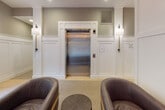 Located at 50 Barnum Ave in Port Jefferson, NY, this elegantly appointed lobby makes a strong first impression with its thoughtfully curated design and upscale finishes. The single-floor space welcomes visitors through large glass entry doors framed by floor-to-ceiling windows that flood the interior with natural light and offer views of the surrounding greenery.

The interior design blends classic and contemporary elements seamlessly. White wainscoting panels line the lower walls throughout, complemented by soft sage-toned upper walls and crisp crown molding. Recessed lighting works in concert with statement brass chandeliers featuring cylindrical glass accents, casting a warm, polished glow across the space. Matching brass wall sconces add a refined continuity to the overall aesthetic.

Comfortable dark leather armchairs with gold-toned legs are arranged in conversational groupings atop neutral carpeting and patterned area rugs, creating inviting seating areas for guests. A stainless steel elevator, framed by white millwork and flanked by wall sconces, serves as a central architectural focal point. Additional corridors lead to mailbox areas and further interior spaces. Light tile flooring in transitional zones ties the design together, resulting in a lobby that is both functional and visually distinguished.