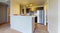 This single-floor residence offers a thoughtfully designed layout that balances functional living spaces with tasteful architectural details. The kitchen is a standout feature, equipped with a full suite of stainless steel appliances including a refrigerator, electric range, and over-the-range microwave. Dark espresso cabinetry contrasts with light quartz countertops, and a white breakfast bar peninsula separates the kitchen from the adjoining living area. Track lighting overhead provides focused illumination throughout the kitchen space, while wood-look flooring adds warmth underfoot.

The living area is highlighted by a white-mantled fireplace and a distinctive arched entryway flanked by decorative columns, adding architectural character to the space. Crown molding runs throughout, lending a refined finish to the neutral-toned walls. Plush carpeting covers the living and bedroom areas, contributing to a comfortable atmosphere. Bedrooms receive natural light through large windows fitted with horizontal blinds, offering views of surrounding greenery.

The bathroom features the same dark cabinetry and light quartz countertop seen in the kitchen, paired with a full tub-shower combination with white tile surround. Wood-look flooring continues here, tying the aesthetic together cohesively throughout the unit.