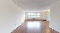 This single-floor residence offers a well-organized layout with distinct living, sleeping, and kitchen zones. The spacious living room is a standout feature, showcasing gleaming hardwood floors, neutral cream-toned walls, and a wide multi-pane window fitted with blinds that allows natural light to fill the room. A wall-mounted air conditioning unit and baseboard heating system provide year-round climate control.

Adjacent to the living room, the galley-style kitchen is efficiently designed with rich dark wood cabinetry, granite-look countertops, and a full suite of stainless steel appliances including a gas range and refrigerator. Light tile flooring completes the functional space. A pendant light fixture illuminates the open pass-through area connecting the kitchen to the living room.

The bedrooms are carpeted and generously sized, each featuring neutral walls, windows with natural light, and baseboard heating. One bedroom includes two closets, offering practical storage. The bathroom features classic tile wainscoting in warm terracotta tones, a pedestal-style sink, and a shower-tub combination. Overall, the space presents a clean, functional interior with a neutral palette throughout.