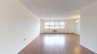 This single-floor residence offers a well-organized layout with distinct living, sleeping, and kitchen zones. The spacious living room is a standout feature, showcasing gleaming hardwood floors, neutral cream-toned walls, and a wide multi-pane window fitted with blinds that allows natural light to fill the room. A wall-mounted air conditioning unit and baseboard heating system provide year-round climate control.

Adjacent to the living room, the galley-style kitchen is efficiently designed with rich dark wood cabinetry, granite-look countertops, and a full suite of stainless steel appliances including a gas range and refrigerator. Light tile flooring completes the functional space. A pendant light fixture illuminates the open pass-through area connecting the kitchen to the living room.

The bedrooms are carpeted and generously sized, each featuring neutral walls, windows with natural light, and baseboard heating. One bedroom includes two closets, offering practical storage. The bathroom features classic tile wainscoting in warm terracotta tones, a pedestal-style sink, and a shower-tub combination. Overall, the space presents a clean, functional interior with a neutral palette throughout.