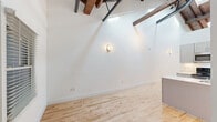 Located at 230 Goldsboro St S in Wilson, NC, this single-floor unit offers a distinctive blend of industrial character and modern updates. The open living area immediately draws the eye upward to dramatic vaulted ceilings adorned with exposed rustic wooden beams and dark metal structural elements, creating a loft-like aesthetic with a sense of generous vertical space. Light-toned hardwood floors run throughout, enhancing the bright, airy atmosphere created by crisp white walls and natural light filtering through windows framed by painted white brick — a nod to the building's historic roots.

The kitchen is well-appointed with gray shaker-style cabinetry, stainless steel appliances including a range with microwave above and a full-size refrigerator, white countertops, and an undermount sink — offering a clean, contemporary cooking space. The galley-style layout maximizes efficiency within a compact footprint.

The bathroom features a full tub-and-shower combination, a white vanity, and a charming penny-tile mosaic floor with gold-toned hardware accents, adding a timeless touch. A dedicated closet provides practical storage. The overall ambiance of the space is one of character-rich, thoughtfully updated living that balances historic architectural details with modern finishes.