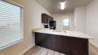 This single-story space is designed with a clean, contemporary aesthetic and a practical open-concept floor plan. The main living area features wood-look luxury vinyl plank flooring in a warm neutral tone, providing a cohesive and durable surface throughout. Natural light filters in through windows fitted with white horizontal blinds, contributing to the bright and airy atmosphere.

The kitchen is a standout feature, equipped with rich dark espresso-toned cabinetry, light granite-patterned countertops, and black appliances including an electric range, over-the-range microwave, and refrigerator. A peninsula with a built-in sink and extended countertop separates the kitchen from the living area, offering additional workspace and casual seating potential.

The entryway provides a welcoming transition into the open living space, with clean wall lines and consistent flooring throughout. The bedroom is carpeted in a neutral gray tone and benefits from two windows that allow ample natural light. It includes a walk-in closet outfitted with double hanging rods and a built-in shelf, providing generous storage capacity.

The bathroom features wood-look tile flooring, a dark vanity with granite-style countertop, a large mirror, and a lighted vanity bar, complemented by neutral wall tones for a polished, cohesive finish.