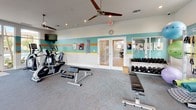 This fitness center offers a comprehensive and inviting workout environment designed to accommodate a wide range of exercise routines. The space is characterized by its distinctive coastal-inspired color scheme, featuring bold teal, orange, and yellow horizontal stripes along the walls, complementing the light, airy atmosphere throughout.

Large windows wrap around much of the facility, flooding the interior with natural light and providing views of lush tropical landscaping, swaying palm trees, and the surrounding outdoor amenity areas. Ceiling fans are positioned throughout to maintain comfortable airflow, and recessed lighting ensures the space remains well-lit at all times.

The cardio zone is equipped with multiple treadmills and elliptical trainers, all positioned to take advantage of the expansive outdoor views. A wall-mounted flat-screen television is conveniently placed nearby for entertainment during workouts. The strength training area features a cable machine system, a multi-station weight machine, and a selection of benches for a variety of exercises. A full dumbbell rack provides an organized and accessible free weights station. Medicine balls, stability balls, and additional functional training accessories round out the equipment offerings.

The flooring transitions between durable rubber and carpet surfaces suited to different workout zones. Glass doors provide direct access to the outdoor pool and patio area, creating a seamless connection between indoor fitness and outdoor recreation.