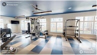Located at 3830 NC-55 E in Durham, NC, this fitness center offers a spacious, single-floor layout designed to support a comprehensive workout experience. The room's most striking design element is the bold navy blue ceiling, which contrasts sharply with the crisp white walls and creates a dynamic, energizing atmosphere. Underfoot, a large-scale blue and tan checkered resilient tile floor adds a distinctive visual character while providing durability and easy maintenance.

Natural light pours in through multiple large windows along the walls, offering views of the surrounding community and keeping the space feeling open and inviting. Overhead, ceiling fans paired with sleek linear LED lighting fixtures ensure the room remains comfortable and well-illuminated throughout the day.

The fitness center is comprehensively equipped for a full range of workouts. A row of cardio machines — including treadmills, ellipticals, a stair climber, and a rowing machine — lines one side of the floor. On the strength training side, a multi-station cable machine, a Smith machine, a dumbbell rack with adjustable bench, medicine balls, kettlebells, and a punching bag provide extensive options for resistance training. A wall-mounted mirror near the free weights area enhances the sense of space, while a water fountain station near the entrance ensures convenient hydration. Wall-mounted TVs are positioned throughout for entertainment during workouts.