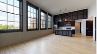 Located at 215 N Webster Ave in Green Bay, Wisconsin, this single-floor unit delivers a compelling blend of industrial character and modern design. The open-concept living and kitchen area immediately impresses with its soaring ceilings and a bank of oversized, black-framed windows that flood the space with natural light while framing views of the surrounding brick streetscape.

The kitchen is a standout feature, outfitted with rich dark cabinetry, a classic white subway tile backsplash, light stone countertops, stainless steel appliances, and a central island with pendant lighting overhead. Light hardwood flooring extends throughout the main living areas, complementing the bold dark trim and door frames that define the unit's aesthetic.

The bedroom areas offer generous proportions, with one room featuring soft carpeting and black-framed windows providing natural light and neighborhood views. Soaring ceiling heights continue into the sleeping quarters, reinforcing the spacious feel. Closet space is accommodated by large double doors in the primary bedroom area.

The bathrooms are thoughtfully designed with classic penny-round tile flooring, white subway tile wainscoting, dark vanity cabinetry with stone-topped sinks, and industrial-style cage lighting fixtures, creating a cohesive, urban aesthetic throughout.