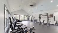 Located at 200 Citygreen Way in Chattanooga, Tennessee, this well-appointed fitness center offers a comprehensive exercise environment within a single, open-plan floor. The space is defined by striking vaulted ceilings that lend an airy, spacious feel, complemented by recessed lighting and a ceiling fan that ensure comfort during workouts.

Large, dark-framed windows line the walls, flooding the interior with natural light and providing pleasant views of the surrounding greenery and community. The durable dark rubber flooring throughout provides both safety and functionality for a wide range of exercise activities.

The facility is equipped with a diverse array of fitness equipment, including treadmills, stationary cycling bikes with digital displays, an elliptical trainer, a multi-station cable and strength machine, a squat rack with bench press, a leg press machine, and various free weights. Stability balls and additional accessories are neatly stored along one wall for versatile training options.

A built-in refreshment and amenity station features grey cabinetry, a countertop with a sink, and a compact refrigerator, allowing users to conveniently hydrate and refresh. Wall-mounted mirrors and posted fitness center rules round out the thoughtfully designed space, making this facility a well-equipped destination for residents seeking a complete workout experience.