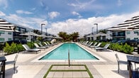 This expansive outdoor amenity space is designed with a contemporary, resort-inspired aesthetic that invites relaxation and social gathering. At its centerpiece is a generously sized rectangular pool with crystal-clear turquoise water, flanked on both sides by a wide sun deck lined with sleek black lounge chairs and striped black-and-white market umbrellas that provide ample shade throughout the day. The clean, large-format tile pavers and accent strips of artificial turf create a polished, modern ground plane that ties the entire area together. Tall palm trees interspersed throughout the deck add a lush tropical character and frame the open sky above. A covered outdoor BBQ station with a stainless steel grill sits beneath a modern pergola-style canopy with dark steel framing, positioned near a gated entry point and overlooking a grassy lawn area suitable for outdoor activities. Adjacent to the main building, a covered colonnade patio offers additional seating with black metal tables and chairs, sheltered beneath a white stucco overhang with large glass doors providing a seamless transition between indoor and outdoor spaces. The surrounding multi-story residential buildings in a crisp white and gray modern palette complete the architectural context, creating a cohesive and well-appointed community outdoor environment.