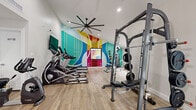Located in Scottsdale, Arizona, this fitness center is a thoughtfully designed workout space that balances energetic aesthetics with a comprehensive selection of exercise equipment. The single-floor layout offers a comfortable footprint, providing enough room to accommodate multiple workout zones without feeling overcrowded.

The space is immediately distinguished by its striking, large-scale abstract mural, which covers an entire accent wall in vivid shades of teal, yellow, orange, blue, and magenta. This bold artistic feature adds a dynamic and motivating atmosphere to the room. The remaining walls are crisp white, keeping the space bright and open, while light wood-tone flooring runs throughout, contributing warmth underfoot.

A large-blade ceiling fan is centrally mounted, ensuring excellent air circulation during workouts. Recessed lighting provides even illumination throughout. A wall-mounted flat-screen TV is positioned for easy viewing from cardio equipment.

The cardio section includes multiple elliptical trainers and a recumbent bike, while the strength training area features a multi-function cable machine, a smith machine with bench press, and a dumbbell rack alongside kettlebells and medicine balls. TRX suspension straps and stability balls round out the functional fitness offerings. Large windows and glass-panel doors allow natural light to filter in, enhancing the overall brightness and openness of the space.