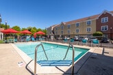 This outdoor pool area is a well-appointed recreational space designed for both relaxation and accessibility. The centerpiece is a generously sized swimming pool with clear, blue-green water, framed by a wide concrete pool deck that provides plenty of room for lounging and movement around the perimeter. A wrought iron security fence encloses the entire area, offering safety and privacy while maintaining an open, airy feel under the expansive open sky.

The deck is furnished with teal lounge chairs and seating clusters shaded by vibrant red patio umbrellas, creating comfortable spots for guests to unwind. A standout feature of the space is its commitment to accessibility — a pool lift is stationed near the entry, ensuring ease of access for all users. The pool area is anchored by a classic brick pool house with a pitched shingle roof, white-trimmed windows, and a covered entryway. Posted pool rules and safety signage are prominently displayed on the exterior brick wall.

The surrounding multi-story buildings feature a mix of beige siding and brick exteriors with traditional window arrangements, contributing to a cohesive, welcoming atmosphere. Mature trees visible beyond the fence add a natural backdrop, completing this thoughtfully designed outdoor retreat.