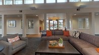 This single-floor communal gathering space is defined by its dramatic architectural character and warm, welcoming ambiance. The centerpiece of the main lounge is a soaring vaulted ceiling clad in warm wood planking, supported by exposed timber beams that lend a lodge-inspired aesthetic to the interior. Clerestory windows line the upper walls throughout, drawing in abundant natural light and offering treetop views that connect the interior to the surrounding landscape.

A stacked-stone fireplace with a light wood mantel anchors one end of the space, providing a cozy focal point and transitioning seamlessly into a built-in seating surround. The main gathering area is generously proportioned, furnished with a mix of plush sectional sofas, upholstered armchairs in neutral and orange tones, and round accent tables that encourage both relaxed lounging and casual conversation. Framed artwork is thoughtfully displayed along the soft, neutral-toned walls, adding a curated, gallery-like quality.

A large floor-to-ceiling window wall at the far end of the room frames outdoor views and bathes the dining area in daylight, where tables and chairs are arranged for communal use. The open floor plan flows between distinct zones — including a secondary lounge area with hardwood flooring and a reception or service counter — creating a versatile, multi-functional environment throughout.