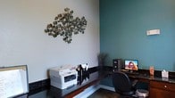 This single-floor office space combines modern aesthetics with practical functionality to create a professional and inviting work environment. The interior design is characterized by a contemporary two-tone color palette, pairing soft neutral walls with a bold teal accent wall that adds depth and visual interest to the space. A striking metal wall sculpture, composed of layered circular disc elements, serves as a focal artistic centerpiece, lending the office a refined and creative character. The workstation area features a sleek, dark granite-topped L-shaped desk that provides ample surface area for day-to-day operations. The desk is equipped with essential office tools, including a multifunction printer, desktop computer, and organizational accessories, reflecting a well-outfitted and efficient workspace. A high-backed ergonomic office chair completes the setup, prioritizing comfort during extended work sessions. Subtle decorative touches, such as potted greenery placed in the corner, contribute to a balanced and pleasant atmosphere. The clean lines, polished surfaces, and thoughtful layout of this office space convey a sense of order and professionalism, making it well-suited for client-facing interactions as well as focused administrative work.