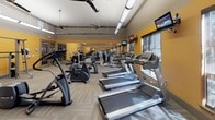 This fitness center is designed to support a comprehensive workout experience within a single, open-plan space. The room is finished with warm yellow accent walls that create an energizing atmosphere, complemented by neutral carpet tile flooring laid in a herringbone pattern for a clean, modern aesthetic. Elevated ceilings with exposed ductwork and industrial-style ceiling fans promote airflow throughout the space, while large windows along one wall invite ample natural light and provide outdoor views.

The cardio zone is generously appointed with multiple treadmills, elliptical trainers, and a stationary bike, each positioned to face wall-mounted flat-screen televisions for an engaging workout experience. The strength training area includes a multi-station cable machine, a Smith machine loaded with weight plates, a dedicated free weight section with a full rack of dumbbells in graduated sizes, and a flat weight bench. Floor-to-ceiling mirrors line one wall, visually expanding the space and allowing users to monitor their form during exercise.

Access to the fitness center is provided through glass-paned French doors, contributing to the open and welcoming character of the space. The overall layout is thoughtfully organized, allowing users to move freely between cardio and strength zones without congestion, making this a functional and well-rounded facility.