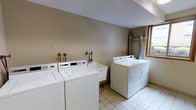 This single-floor laundry facility is a practical, well-equipped space designed to meet shared laundry needs. The room features multiple top-loading washing machines and dryers arranged along the walls, providing ample capacity for simultaneous loads. A built-in utility sink with exposed plumbing fixtures is conveniently positioned between the washers, offering added functionality for hand-washing or pre-treating garments. Ventilation ductwork is visibly routed along the back wall, ensuring proper airflow and safe operation of the dryers. The space is finished with neutral beige walls and durable tile flooring, creating a clean and easy-to-maintain environment. A window on one side of the room allows for natural light to enter, contributing to a brighter and more pleasant atmosphere. The layout is straightforward and efficient, with machines positioned to allow comfortable access and movement throughout the room. Overall, this laundry facility is a no-frills, utilitarian space that prioritizes function and convenience, making it a reliable amenity for its users.