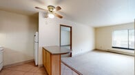 This single-floor unit offers a straightforward, functional layout designed for comfortable everyday living. The kitchen is equipped with warm oak cabinetry, laminate countertops, a stainless steel sink, an electric range with overhead ventilation, and a full-size refrigerator. Tile flooring runs through the kitchen area, which flows into the adjoining living space via a peninsula-style counter with additional cabinet storage beneath — a practical divide that maintains an open connection between the two areas. A ceiling fan with a light fixture overhead serves the living area, which transitions to wall-to-wall carpeting. The bedroom areas are carpeted and each features a closet with a hanging rod for storage. One bedroom also benefits from a window with natural light and a baseboard heater. The bathroom includes a full tub and shower combination with classic tile surround in neutral tones, paired with standard fixtures. Throughout the unit, neutral wall colors and light finishes create a clean, understated aesthetic. The layout is compact and efficient, making good use of the available space across each room.