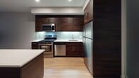 This single-floor unit showcases a thoughtfully designed layout with a consistent modern aesthetic carried throughout each space. The kitchen serves as a standout feature, boasting rich, dark wood cabinetry that extends to the ceiling for ample storage, crisp white countertops, and a full suite of stainless steel appliances including a gas range, built-in microwave, dishwasher, and refrigerator. Subtle under-cabinet lighting illuminates a glass tile backsplash, adding a refined touch to the culinary space. A kitchen island with a contrasting dark base and white surface provides additional prep and serving space.

Light hardwood flooring flows through the main living areas, lending warmth and continuity to the open layout. A long, well-lit hallway connects the living spaces to the private areas of the unit, lined with clean white walls and featuring a louvered storage closet for added functionality. Recessed lighting throughout ensures even, practical illumination.

The bathroom is finished with light-toned tile flooring and walls, featuring a soaking tub and shower combination with bright white square tile surround and brushed nickel fixtures. The overall palette of the unit is neutral and understated, with light gray walls and white trim creating a clean, move-in-ready environment suited to a variety of interior styles.