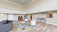 Located at 191 Spencer St in Manchester, Connecticut, this single-floor community amenity space offers a generously sized, open layout designed for both relaxation and recreation. The space is defined by its dramatically high ceilings, which are accentuated by elegant crown molding and a two-tone wall treatment that adds architectural depth and visual interest throughout.

Light wood-look flooring flows seamlessly across the entire space, contributing to a clean, contemporary aesthetic. A welcoming lounge area anchors the interior, furnished with white barrel chairs arranged around a geometric-patterned area rug in gray, blue, and yellow — adding a vibrant focal point to the room. A coffee and refreshment station, along with a small literature and amenity display, lines one wall, enhancing the space's functionality.

Floor-to-ceiling windows along the entry corridor flood the space with abundant natural light and offer views of the surrounding outdoor environment. A dedicated game area features a full-size pool table alongside bar-height seating, making it ideal for social gatherings. The neutral warm-gray wall palette, recessed lighting, and crisp white trim details unify the space with a polished, modern feel. The overall ambiance is open, bright, and inviting — well-suited for community engagement and everyday use.