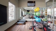 This single-floor yoga and fitness studio, part of the Vida community, is thoughtfully designed to support a wide range of exercise and wellness activities. The space is defined by rich hardwood-style flooring that spans the open main area, providing a clean, durable surface ideal for yoga, stretching, and group fitness classes. A full wall of floor-to-ceiling glass partitions with double-entry doors connects the studio to an adjacent fitness center, creating a sense of openness and visual continuity between the two spaces. Large windows along one wall invite natural light and offer views of mature trees and the surrounding landscape, contributing to a calm, motivating atmosphere. Full-length mirrors on one wall enhance the sense of space while allowing users to monitor their form during workouts. The studio is equipped with stationary cycling bikes, a variety of stability and exercise balls in multiple sizes, step platforms, balance boards, and a rack of free weights, offering versatile options for both group and individual training. Wall-mounted flat-screen televisions are positioned for easy viewing throughout the room. A designated area near the entrance features woven storage baskets and posted room rules, keeping the space organized and community-oriented. Recessed lighting supplements natural light for consistent illumination throughout the day, making this an inviting, fully functional fitness environment.