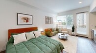 Located at 100 Callan Ave in San Leandro, California, this thoughtfully designed studio unit makes efficient use of its single-floor layout. A welcoming hallway fitted with recessed lighting and a sliding door leads into the open-plan main area, where the living, sleeping, dining, and kitchen functions are seamlessly integrated.

The living and sleeping area is bright and airy, benefiting from a large window that invites natural light and offers a view of the surrounding greenery. Wood-look plank flooring runs throughout, lending warmth and visual continuity to the space.

The kitchen is well-appointed with light wood-grain cabinetry, white quartz countertops, and a soft green subway tile backsplash. Stainless steel appliances — including a refrigerator, range, and over-the-range microwave — provide full functionality within a compact footprint. Open shelving adds character and additional storage.

The full bathroom features a soaking tub and shower combination, built-in shelving for linens, and the same wood-look flooring found throughout the unit, maintaining a cohesive aesthetic. A dedicated closet provides practical storage, while the private patio extends the living space outdoors, offering a personal outdoor retreat.

Clean white walls, recessed lighting, and modern finishes throughout create a bright, contemporary ambiance suited to efficient urban living.